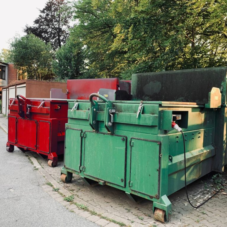 on an industrial site a green waste compactor and a red waste compactor stand side by side