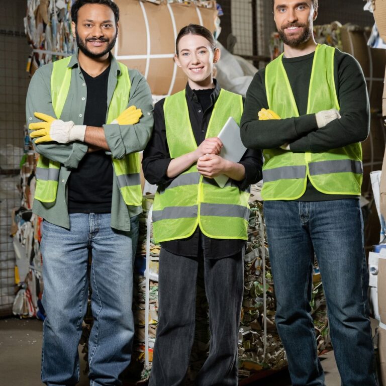worker in protective vest holding digital tablet and standing near multiethnic colleagues and waste paper in waste disposal station